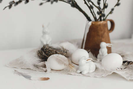 Happy Easter! Natural eggs, bunny figurines, feathers, nest, willow branches in vase on aged wooden table. Easter rustic still life. Simple stylish easter decoration on table.の写真素材