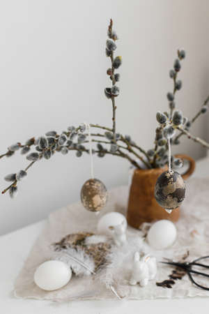 Happy Easter! Natural eggs, bunny figurines, feathers, nest, willow branches in vase on aged wooden table. Easter rustic still life. Simple stylish easter decoration on table.の写真素材