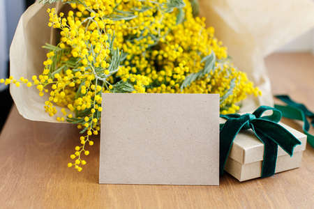 Happy womens day and Happy Mothers day. Stylish mimosa bouquet, gift box and greeting card on wooden background. Greeting card template with space for text. 8 march conceptの写真素材