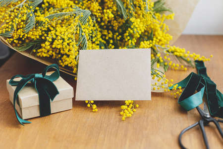 Happy womens day and Happy Mothers day. Stylish mimosa bouquet, gift box and greeting card on wooden background. Greeting card template with space for text. 8 march conceptの写真素材
