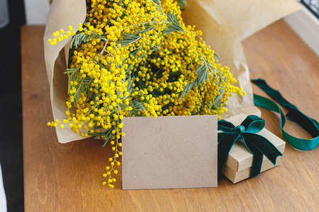 Stylish mimosa bouquet, gift box and greeting card on wooden background. Happy womens day and Happy Mothers day. Greeting card template with space for text. 8 march conceptの写真素材