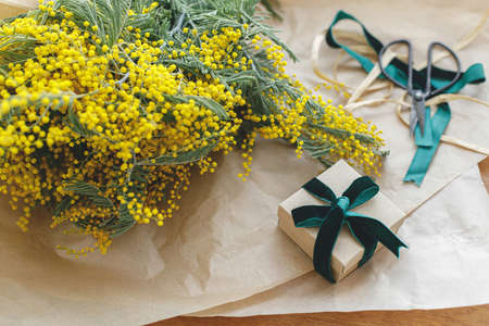 Stylish mimosa bouquet and gift box on wooden background. Happy womens day and Happy Mothers day. Beautiful mimosa flowers in craft paper and gift with green ribbon on windowsill. 8 marchの写真素材