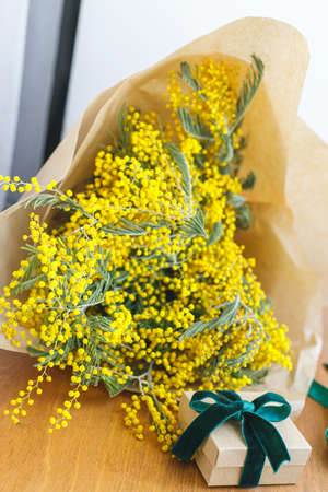 Happy womens day and Happy Mothers day. Stylish mimosa bouquet and gift box on wooden background. Beautiful mimosa flowers in craft paper and gift with green ribbon on windowsill. 8 marchの写真素材