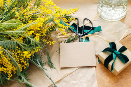 Happy womens day and Happy Mothers day. Stylish mimosa bouquet, gift box and greeting card on wooden background. Beautiful mimosa flowers in craft paper and gift with green ribbon. 8 marchの写真素材