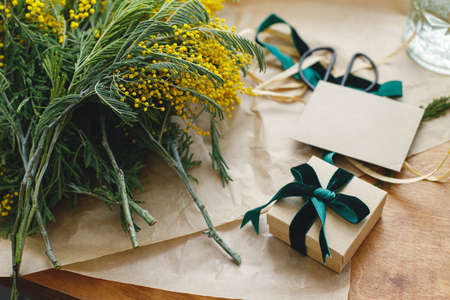 Happy womens day and Happy Mothers day. Stylish mimosa bouquet and gift box on wooden background. Beautiful mimosa flowers in craft paper and gift with green ribbon on windowsill. 8 marchの写真素材