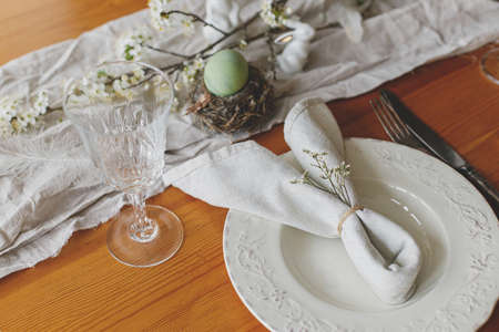 Stylish Easter brunch table setting. Easter egg in nest, bunny napkin on plate, cutlery, bunny,...