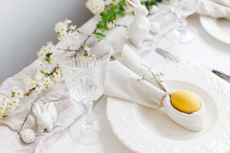 Happy Easter! Stylish elegant Easter brunch table setting. Easter egg in bunny napkin on plate with cutlery, bunny, spring flowers on rustic white table. Modern eco table arrangementの写真素材