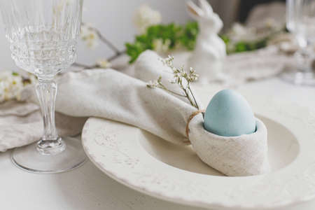 Happy Easter! Stylish elegant Easter brunch table setting. Easter egg in bunny napkin on plate with cutlery, bunny, spring flowers on rustic white table. Modern eco table arrangementの写真素材