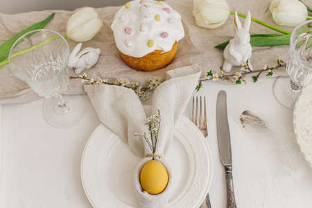 Stylish Easter egg in bunny napkin on plate, modern easter cake, cutlery, bunny, spring flowers on...