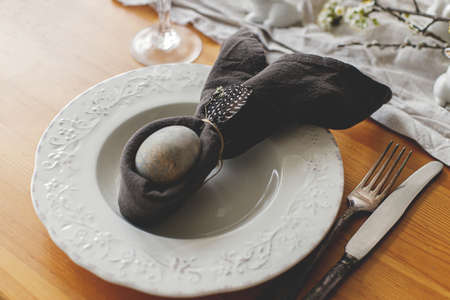 Stylish Easter brunch table setting. Easter egg in bunny napkin on plate with cutlery, bunny,...