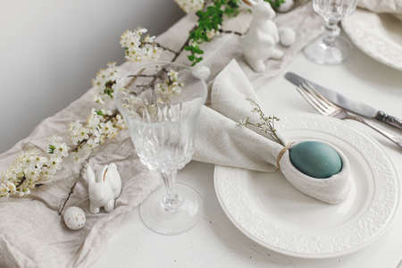 Stylish elegant Easter brunch table setting. Easter egg in bunny napkin on plate with cutlery, bunny, spring flowers on rustic white table. Modern Easter table arrangementの写真素材