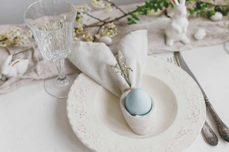 Happy Easter! Stylish elegant Easter brunch table setting. Easter egg in bunny napkin on plate with cutlery, bunny, spring flowers on rustic white table. Modern eco table arrangementの写真素材
