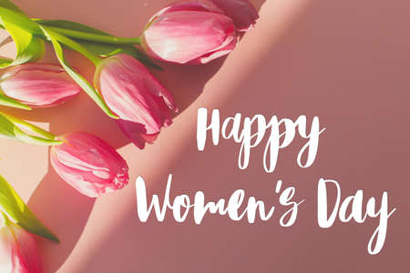 Happy womens day text on pink tulips in sunlight on pink background flat lay. Stylish greeting card....