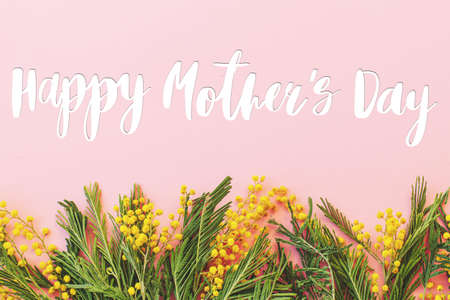 Happy mothers day text on mimosa flowers flat lay on pink background. Stylish greeting card. Happy Mother's Day, gratitude and love to mom. Handwritten letteringの写真素材