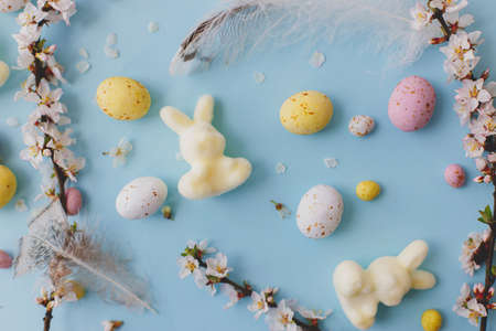 Happy Easter! Colorful Easter chocolate eggs, bunny, cherry blossoms, feathers layout on blue background. Easter flat lay. Greeting card or banner. Stylish tender spring imageの写真素材