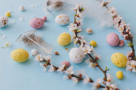 Happy Easter! Colorful Easter chocolate eggs, cherry blossoms, feathers layout on blue background. Easter background. Greeting card or banner. Stylish tender spring imageの写真素材