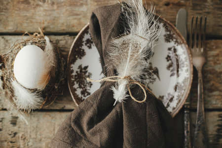 Rustic Easter table setting top view. Natural egg in nest with feathers, vintage plate, cutlery,...