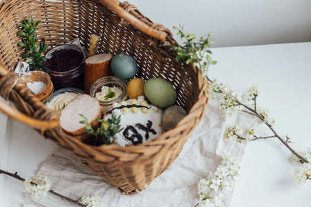 Natural dyed easter eggs, easter bread, ham, beets, butter, cheese in wicker basket decorated with green buxus on rustic wooden table. Traditional Easter basket foodの写真素材