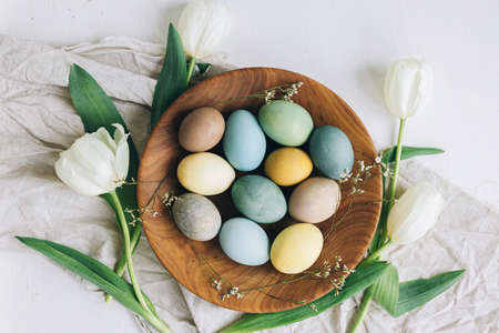 Happy Easter! Stylish Easter eggs in wooden plate, tulips and linen napkin on rustic white table, flat lay. Natural dyed colorful eggs and spring flowers rustic composition.の写真素材