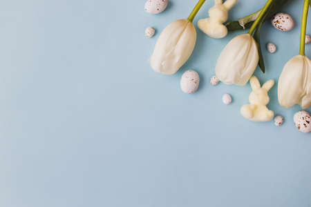 Stylish easter eggs, tulips and bunnies flat lay on blue background. Modern chocolate easter eggs and white flowers layout. Greeting card template, space for text. Happy Easterの写真素材