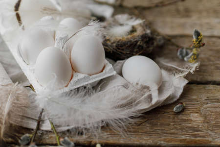 Stylish Easter rustic still life. Easter natural eggs in tray, feathers, willow branches, nest, linen cloth on aged wooden table. Happy Easter! Simple rural aestheticsの写真素材