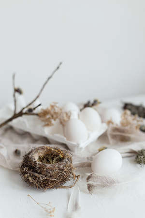 Easter simple aesthetics. Easter natural eggs, feathers, willow branches, nest on rustic white table. Stylish rural Easter still life in pastel white and beige colors.の写真素材