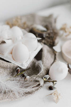 Simple rural Easter aesthetics. Natural eggs in tray, feathers, willow branches, nest on burlap on rustic white wooden table. Happy Easter. Easter rustic still life.の写真素材