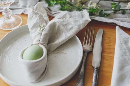 Happy Easter! Easter egg in bunny napkin on plate with cutlery, bunny, spring flowers and rustic cloth on wooden table. Stylish Easter brunch table setting. Eco friendly decorationの写真素材