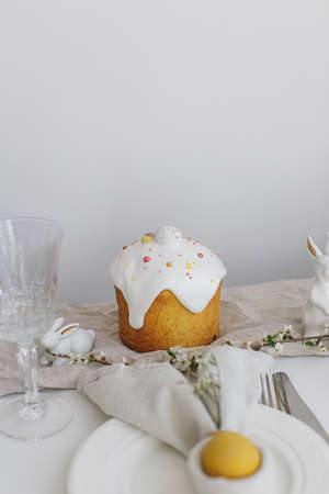 Stylish Easter brunch table setting. Modern Easter cake, egg in bunny napkin on plate, cutlery, bunny, spring flowers on rustic white table. Easter table arrangement and eco friendly decorationsの写真素材