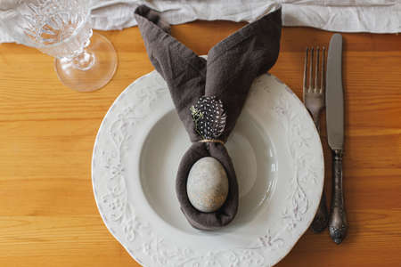 Stylish Easter brunch table setting. Easter egg in bunny napkin on plate with feather, cutlery,...