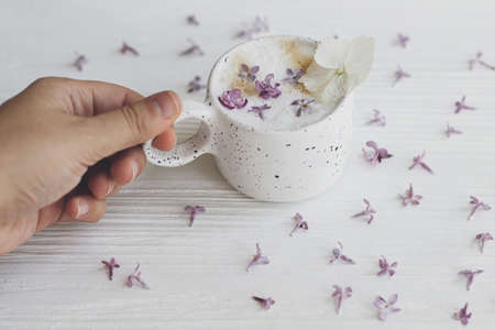 Hand holding delicious coffee with lilac petals on white wooden background. Happy mothers day. Good morning. Beautiful  flowers in stylish cup of coffee.の写真素材