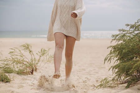 Beautiful woman legs barefoot with sand walking on sandy beach, carefree vacation mood. Stylish young female in knitted sweater relaxing on coast and having fun, cropped viewの写真素材