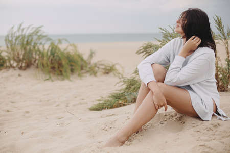 Beautiful woman with windy hair sitting on sandy beach on background of green grass and sea, calm tranquil moment. Stylish young female in sweater relaxing on coastの写真素材