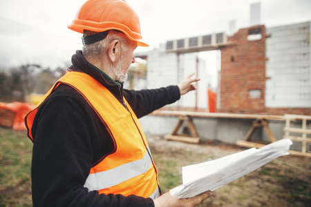 Senior male foreman in hardhat checking blueprints of building new modern house. Man engineer or...
