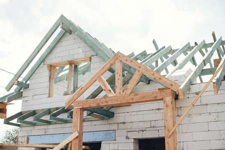 Unfinished modern farmhouse building. Wooden roof framing of mansard with dormer and aerated concrete block walls with windows. Rafters and beams on blocks. New house constructionの写真素材