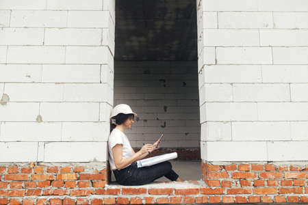 Stylish happy woman engineer in hard hat looking at digital plans on tablet while sitting in window...