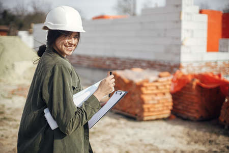 Stylish happy woman architect with blueprints and plans at construction site. Young female engineer or construction worker in hardhat looking at building new modern house. Copy spaceの写真素材
