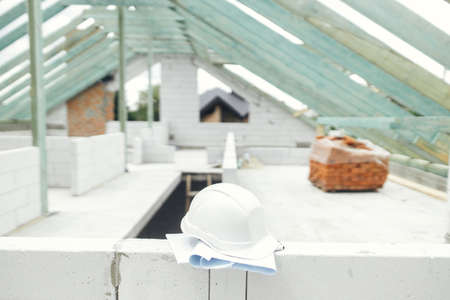 Architecture and House building concept. Hard hat with house blueprints on aerated concrete blocks on background of unfinished wooden roof framing. Safety helmet on construction siteの写真素材
