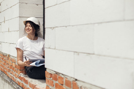 Stylish happy woman engineer in hard hat looking at digital plans on tablet while sitting in window...