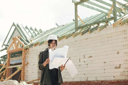 Stylish happy woman architect checking blueprints against modern farmhouse construction site. Young female engineer or construction worker in hardhat with plans of new modern houseの写真素材