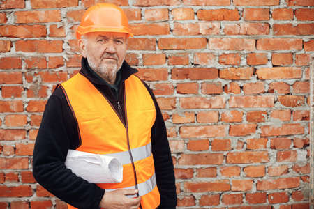 Senior man engineer or construction worker in hardhat standing with blueprints at masonry brick wall at building new house. Portrait of male foreman holding plans at construction siteの写真素材