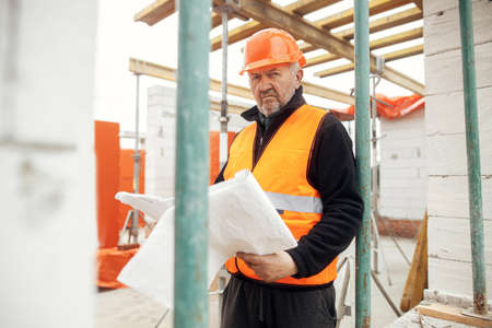 Senior male foreman in hardhat checking blueprints of building new modern house. Man engineer or construction worker looking at plans at construction site. Copy spaceの写真素材