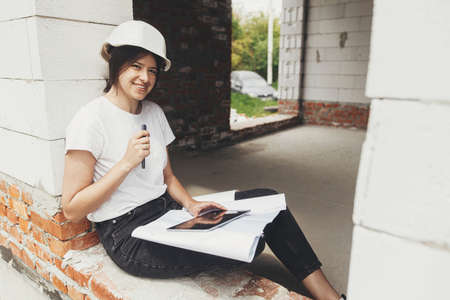 Stylish happy woman engineer in hard hat looking at digital plans on tablet while sitting in window...