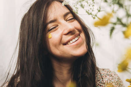 Beautiful happy woman with yellow petals under eyes posing with wildflowers in sunny room. Portrait of young brunette female in boho dress smiling with flowers. Skin care and self care. Summer moodの写真素材