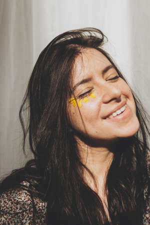 Beautiful happy woman with yellow petals under eyes close up in sunny room. Aging skin care and self care. Summer mood. Portrait of young brunette boho female with flowers.の写真素材