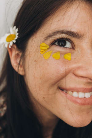 Beautiful happy woman with yellow petals under eyes close up. Portrait of young brunette boho female with daisy flower. Aging skin care and self care. Summer moodの写真素材