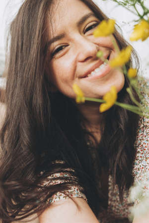 Beautiful happy woman in boho dress posing with wildflowers in sunny room. Portrait of young brunette female smiling with flowers. Skin care and self care. Summer moodの写真素材