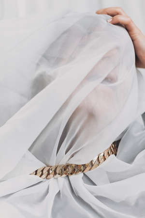 Social isolation and pressure. Woman suffocates under veil, posing with golden necklace. Portrait of young female covered with light fabric. Self care and emotional break conceptの写真素材