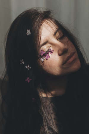 Beautiful sensual woman with lilac petals on face and hair in sunlight. Moody portrait of young brunette with flowers on face in light and shadow. Skin care and natural cosmetics conceptの写真素材
