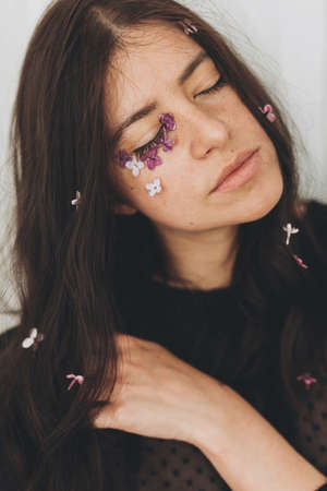 Beautiful sensual woman with lilac petals on face and in hair. Moody portrait of young brunette with flowers on face indoors. Skin care and natural cosmetics concept. Femininity Self careの写真素材